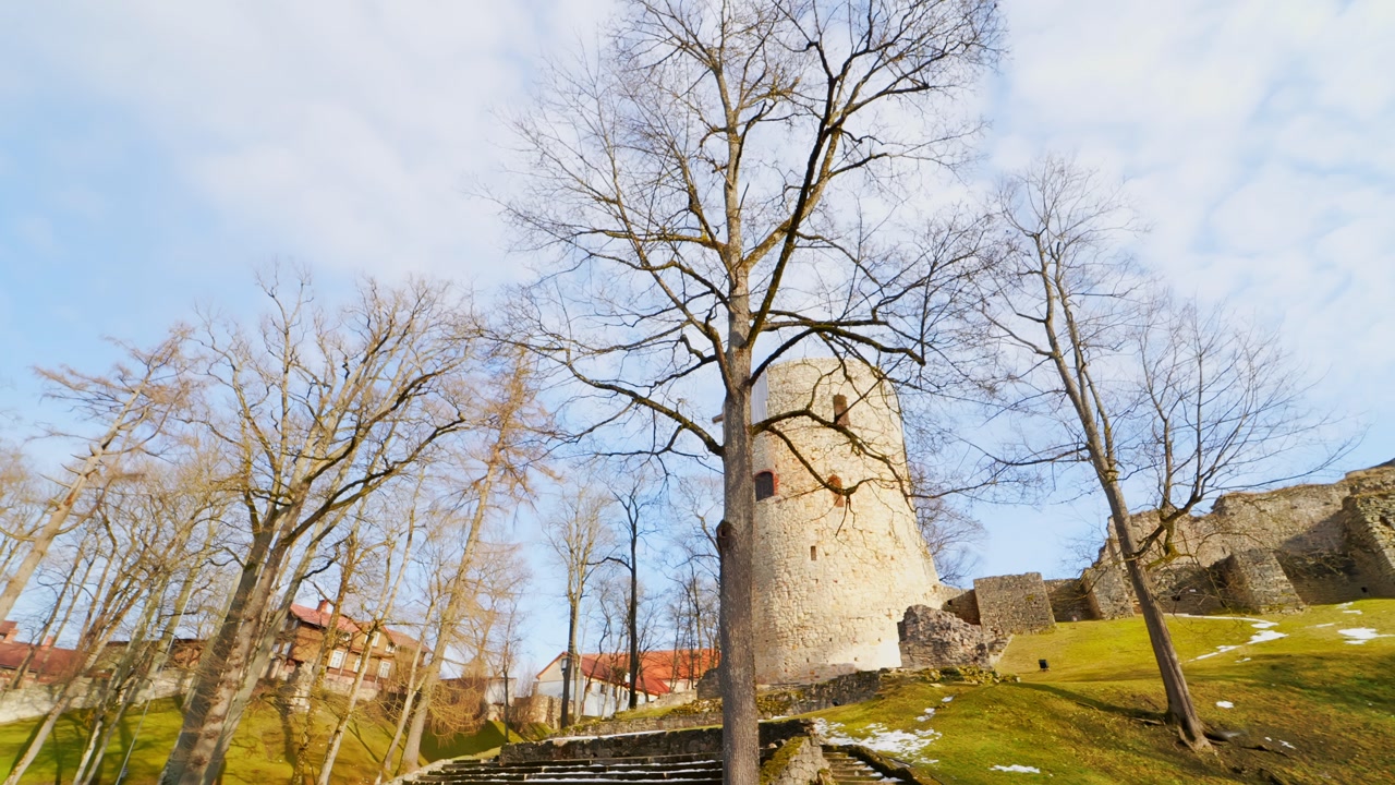 Round medieval tower above stone stairs in Cesis, framed by bare trees and bright spring light, capturing historic fortifications and a peaceful old castle landscape.