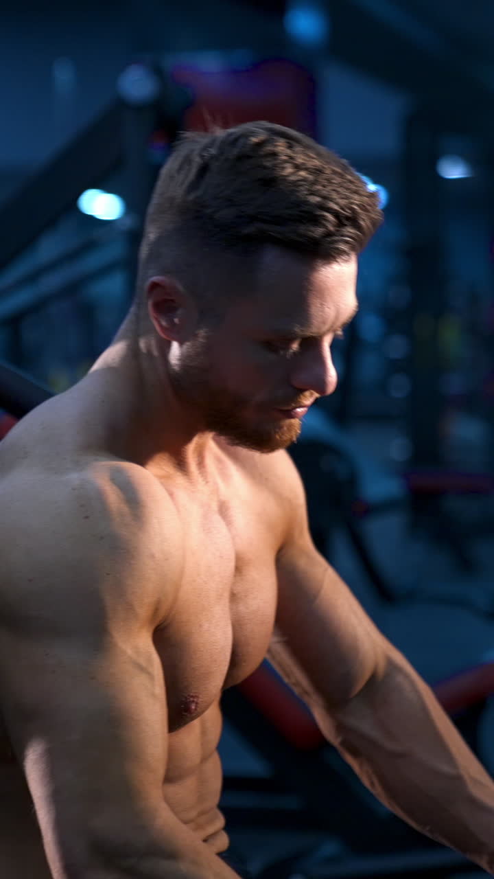 Brutal strong athletic man pumping up muscles. Doing workout on sport equipment. Muscular bodybuilder doing exercises in gym with naked torso. Fitness and bodybuilding concept. Video from the side. Vertical video