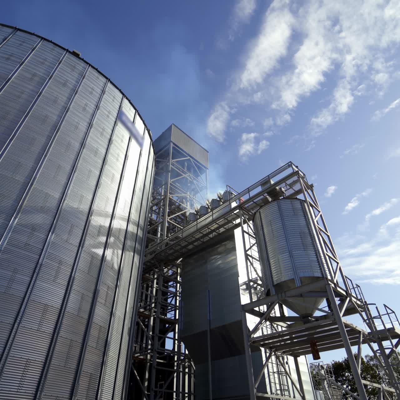 Grain elevators for drying seeds. Modern grain terminal. White smoke goes into the air from grain-drying complex. Metal tanks of elevator outdoors.