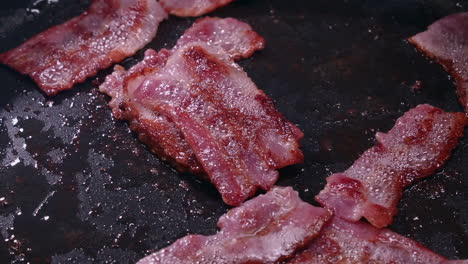 fork stacks sputtering cooked bacon strips in hot saucepan, close-up