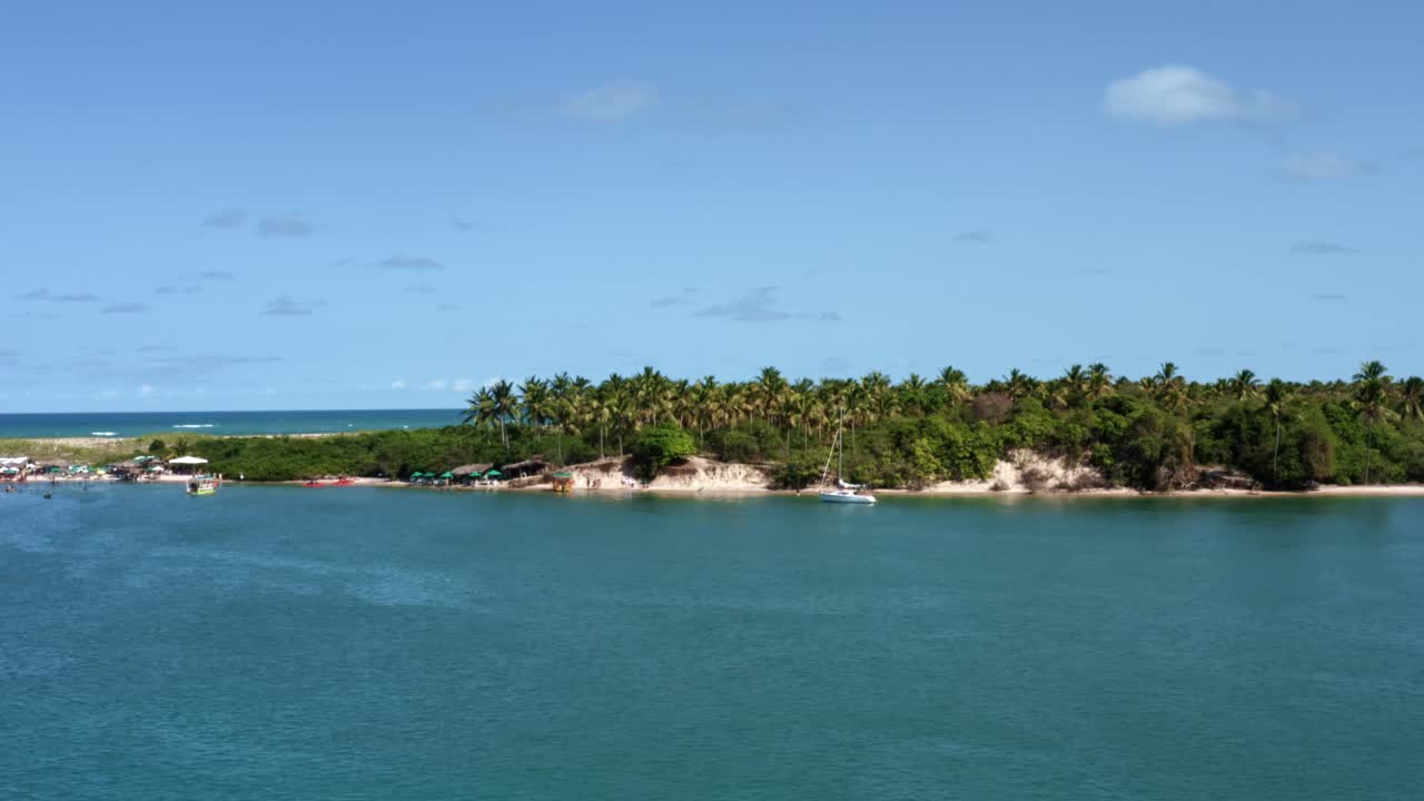 Left trucking aerial drone shot of the beautiful tropical touristy Restinga beach where the large Curimataú river meets the sea near Barra do Cunhaú in Rio Grande do Norte, Brazil on a summer day