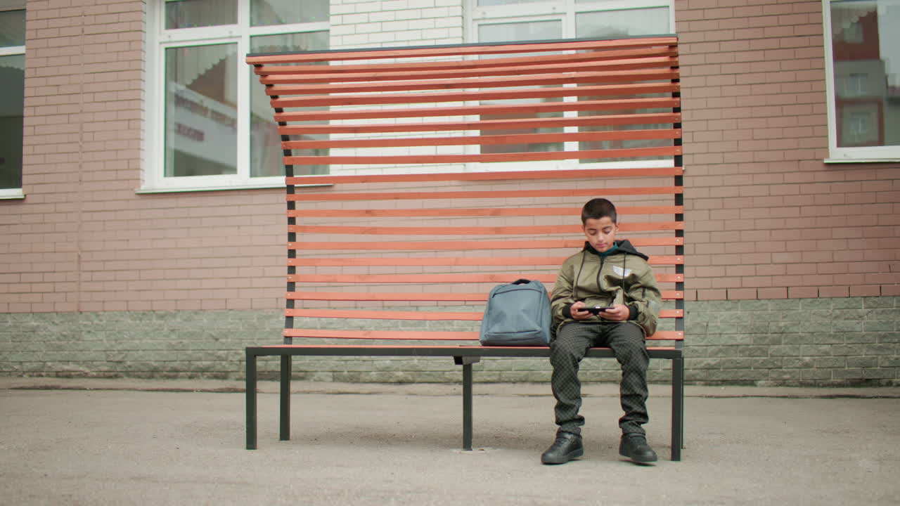 Niño sentado en un banco moderno jugando con el móvil durante el recreo con una mochila a su lado; mira a izquierda y derecha antes de volver a concentrarse en la pantalla; el reflejo de un edificio se ve en la ventana de cristal de una escuela urbana.