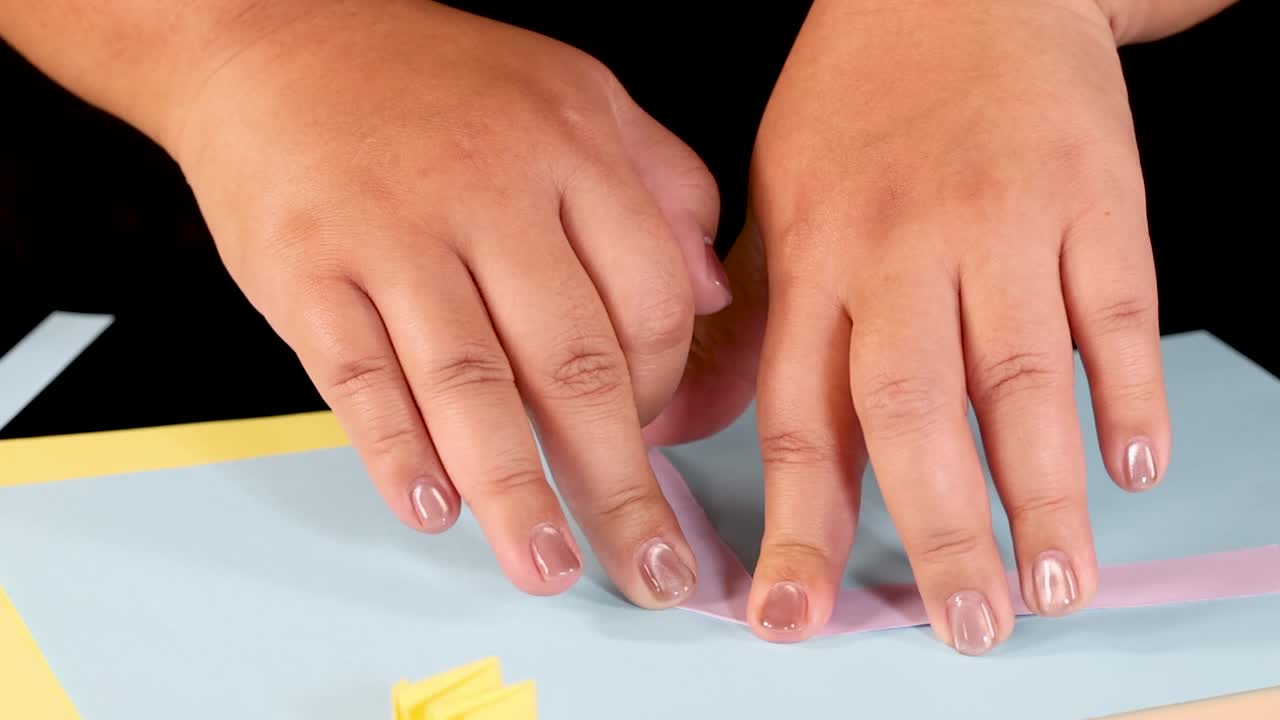 A person's hands carefully fold and crease a thin pink paper strip on blue cardstock