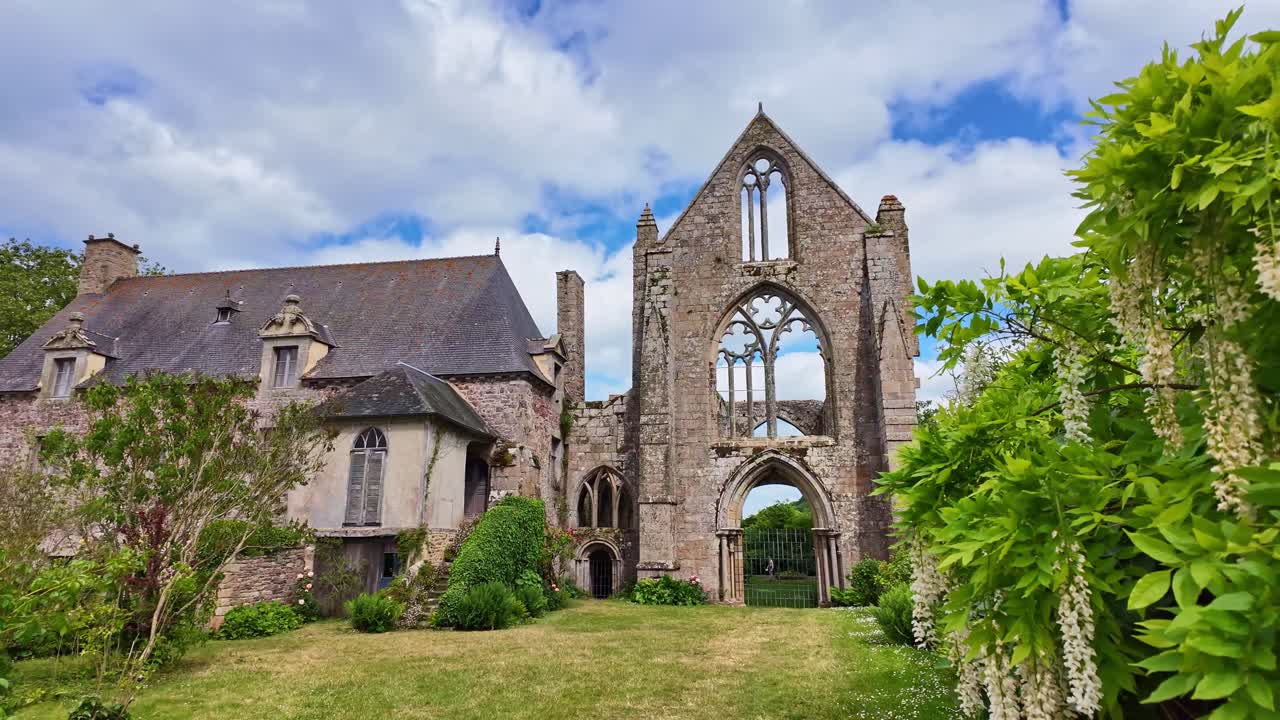 Camera pulls back revealing Beauport Abbey ruins surrounded by flowering trees under a partly cloudy sky