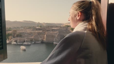 young woman travelling wakes up in a hotel room and stands by the window enjoying the nile in aswan, egypt