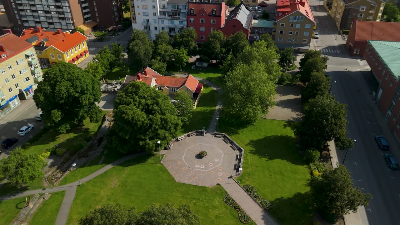 Parallax-kredsløb omkring grønne Betty Backs Park, afslørende centralt monument og frodige træer omgivet af bygader og beboelsesejendomme i Trollhättan under blå himmel.