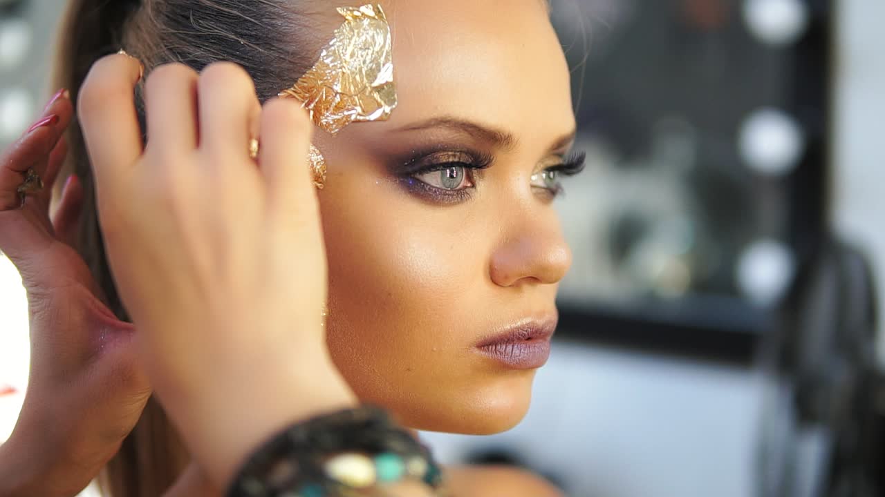 Close up shot of a makeup artist applying golden shiny pieces of metallic paper on a model's face. Preparing for the fashion