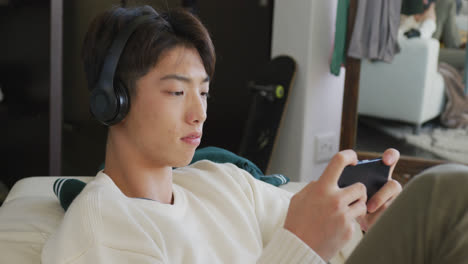 asian boy wearing headphones playing game on smartphone sitting on the couch at home