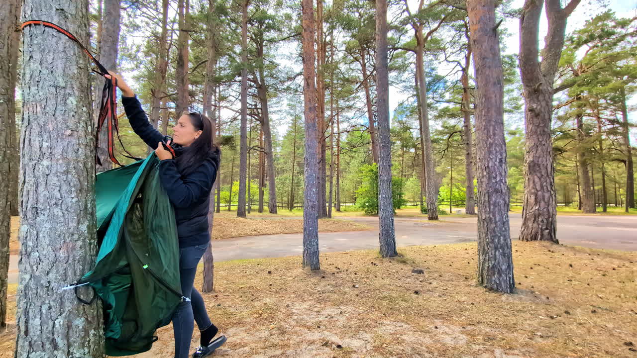 A woman hanging a hammock between trees in a forest park near Engure, Latvia