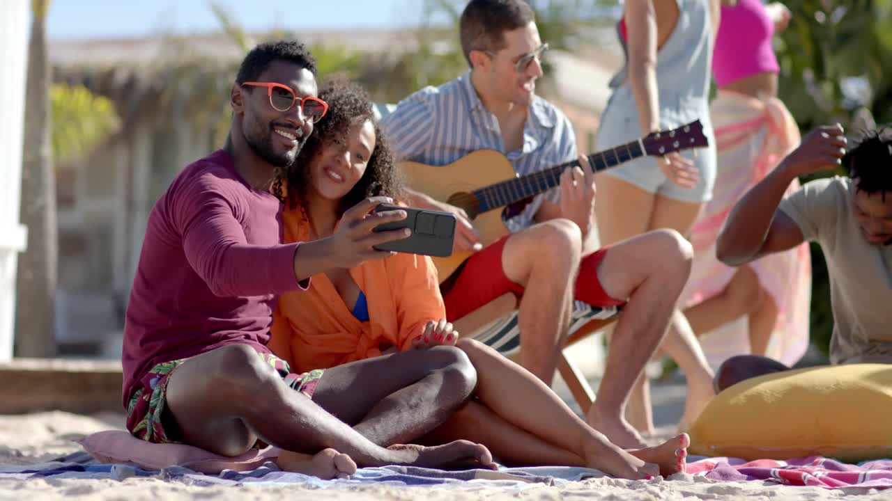 un grupo feliz y diverso de amigos tomando selfies y tocando la guitarra en la playa con la casa de la playa