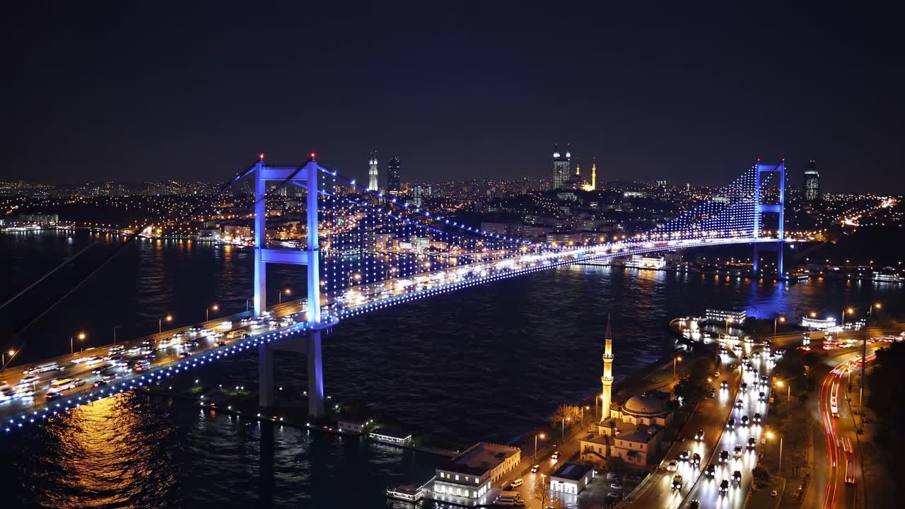 The Bosphorus Bridge in Istanbul, Turkey glowing a vibrant blue at night with car traffic crossing and city lights illuminating the background