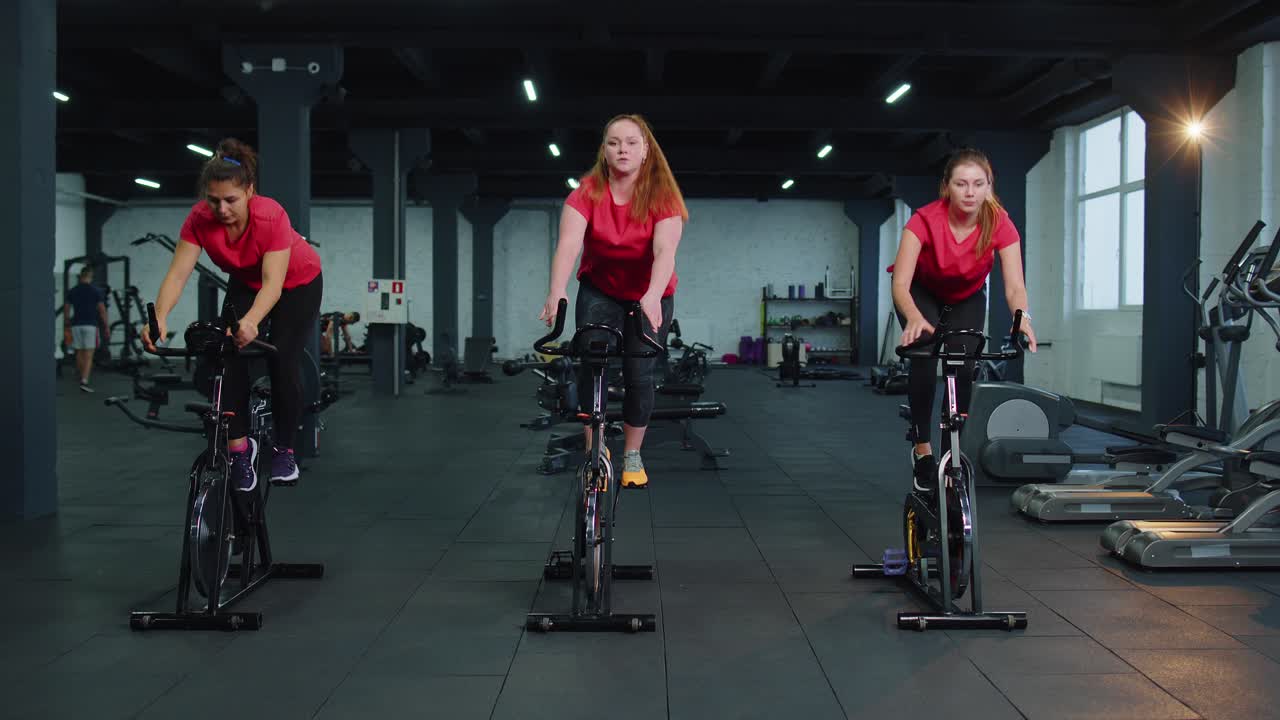 건강한 백인 여성 그룹이 체육관에서 정지 자전거 기계 자전거에서 운동을합니다.
