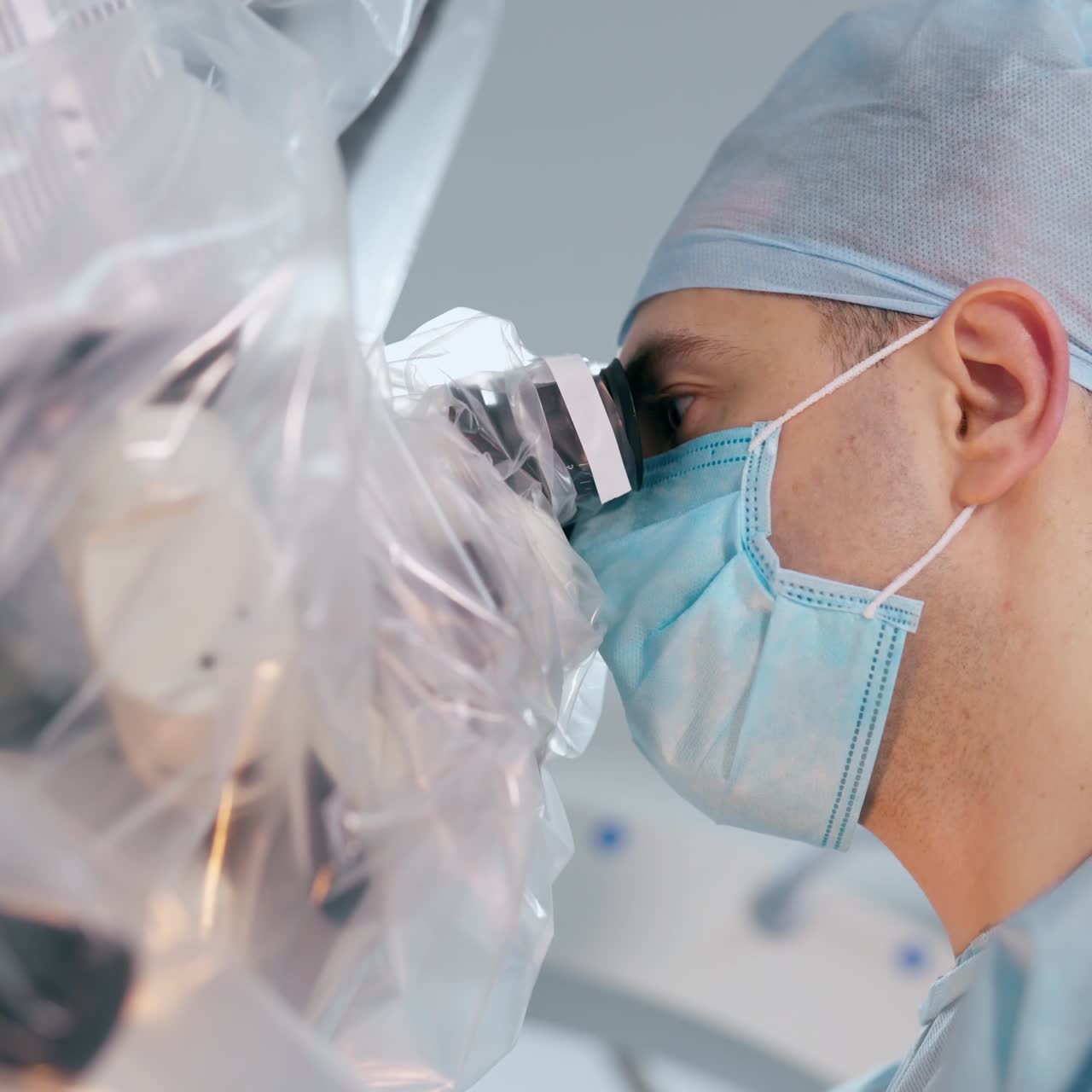Male surgeon in blue mask and cap. Side view of a doctor looking into medical microscope during the surgery. Close-up.