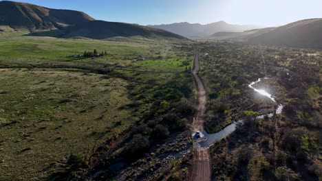 Aerial-shot-of-car-driving-towards-camera-on-dirt-road-in-Willcox,-Arizona,-wide-rotating-drone-shot-with-mountains-in-the-background-and-dust-behind-the-car