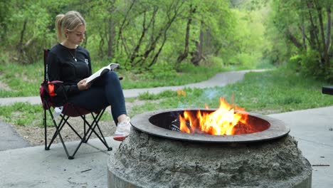 Hermosa-Mujer-Morena-Sentada-Y-Leyendo-Un-Libro-Junto-A-La-Fogata-Durante-El-Día-En-American-Fork-Canyon,-Utah