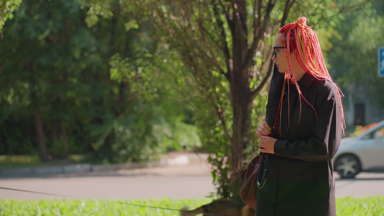 거리에서 다채로운 땋은 머리를 한 여성, 도로 환경 근처에서 생동감 넘치는 머리를 한 사람, 다색 땋은 머리를 하고 캐주얼하게 옷을 입은 여성이 차량이 지나다니는 도시 길가 근처에서 잠시 멈춰 서 있어요.