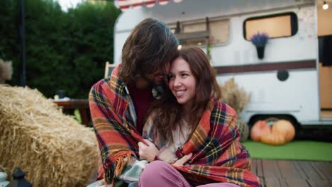 happy brunette guy in a checkered plaid sits wrapped up with his brunette girlfriend during his picnic near a trailer outside the city in a camp in the summer