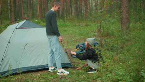 couples setting up tent for camping in forest as man hands peg to lady playfully while scratching neck, both enjoying lighthearted teamwork in green outdoor environment surrounded by tall pine trees