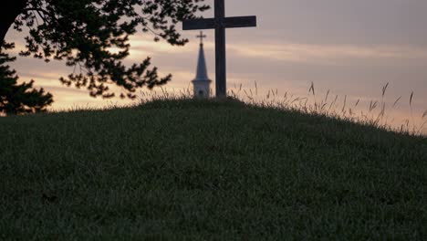 A serene video still of a cross on a grassy hill at sunset, with a church steeple in the background