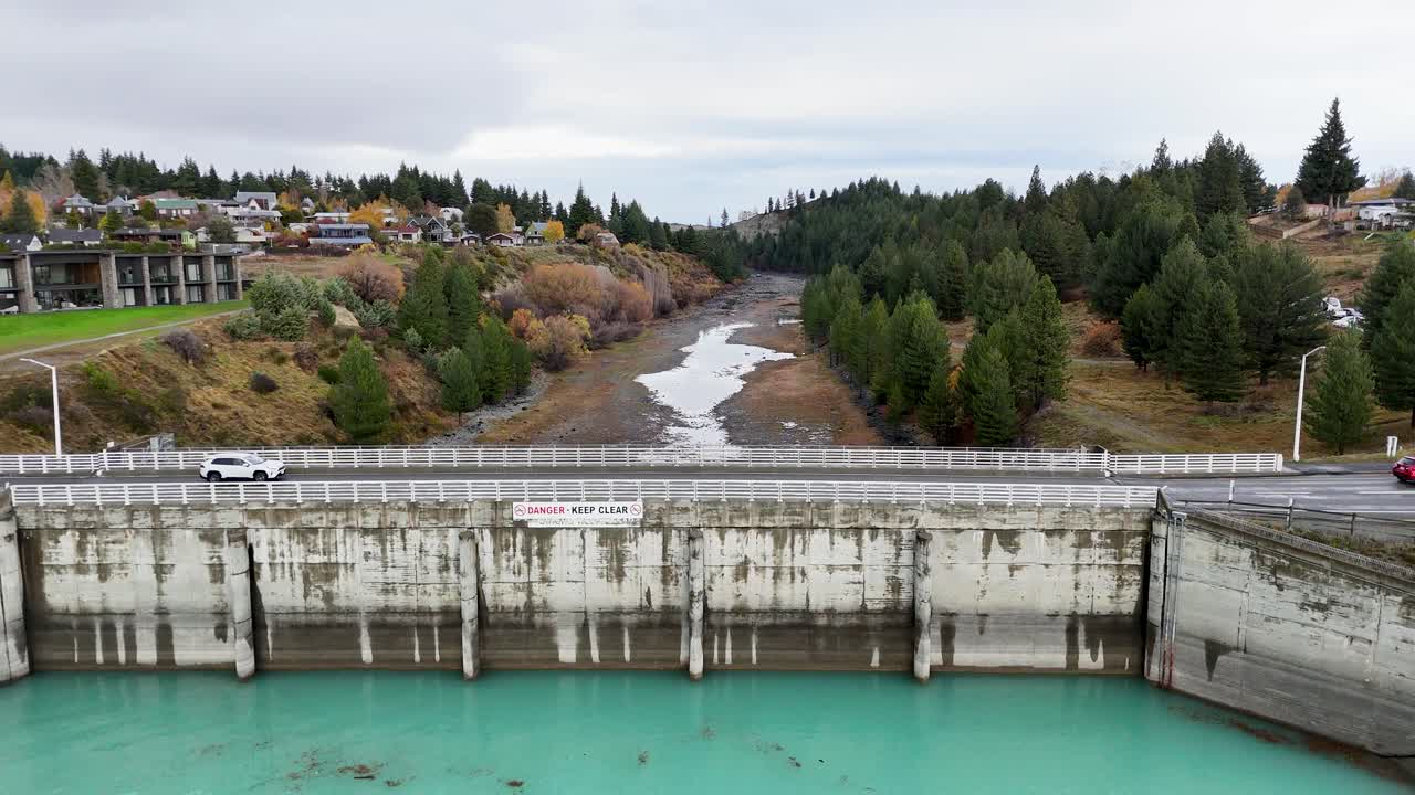 Veicoli attraversano un ponte su acque turchesi al Lago Tekapo, con una vegetazione lussureggiante e cieli coperti sullo sfondo