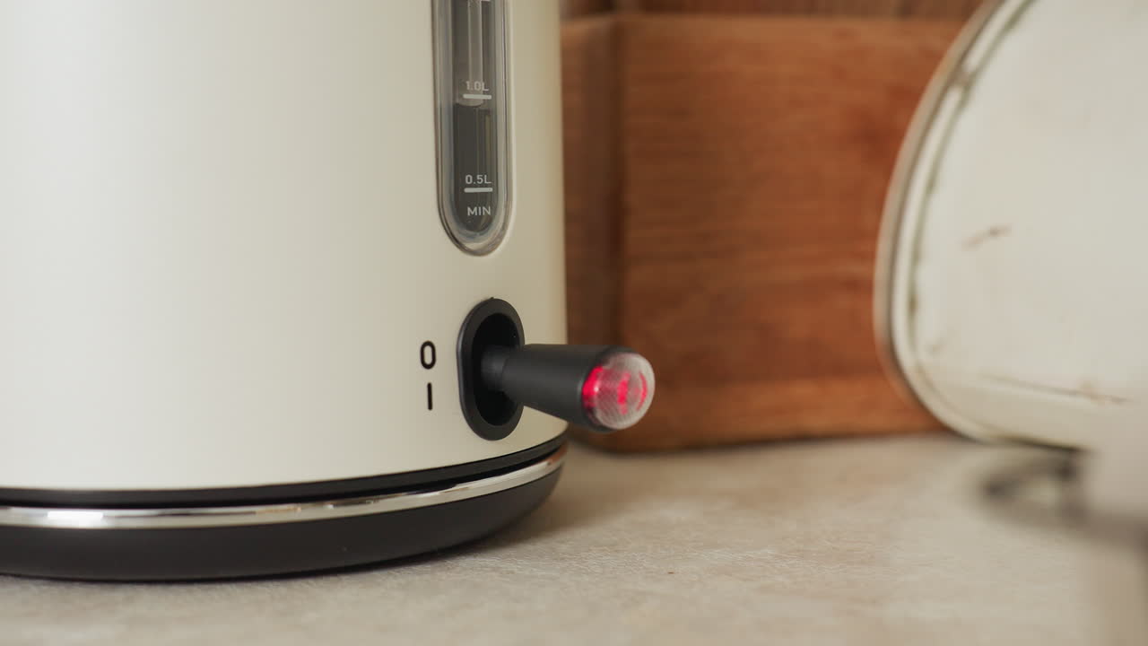 Close up of hand pressing electric kettle switch with indicator light blinking, set against warm-toned kitchen background with wood and metal textures creating a cozy cooking environment