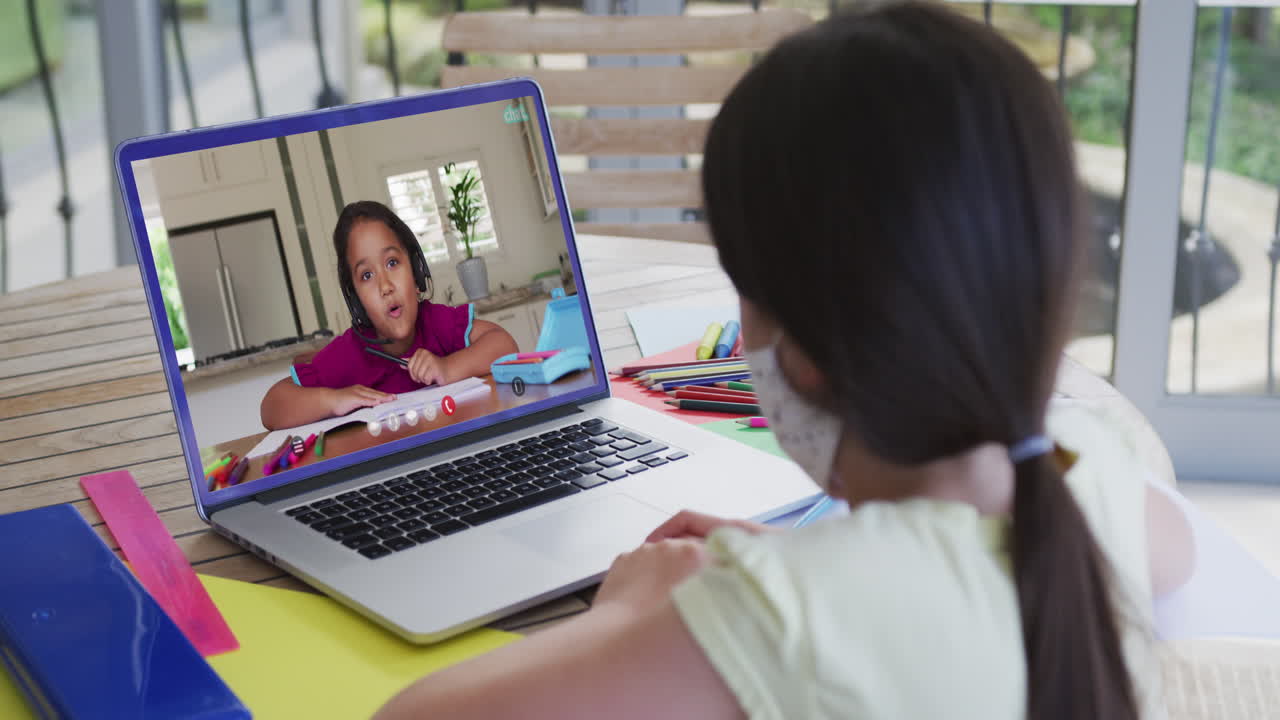 chica biracial sentada en un escritorio usando una computadora portátil teniendo una lección escolar en línea