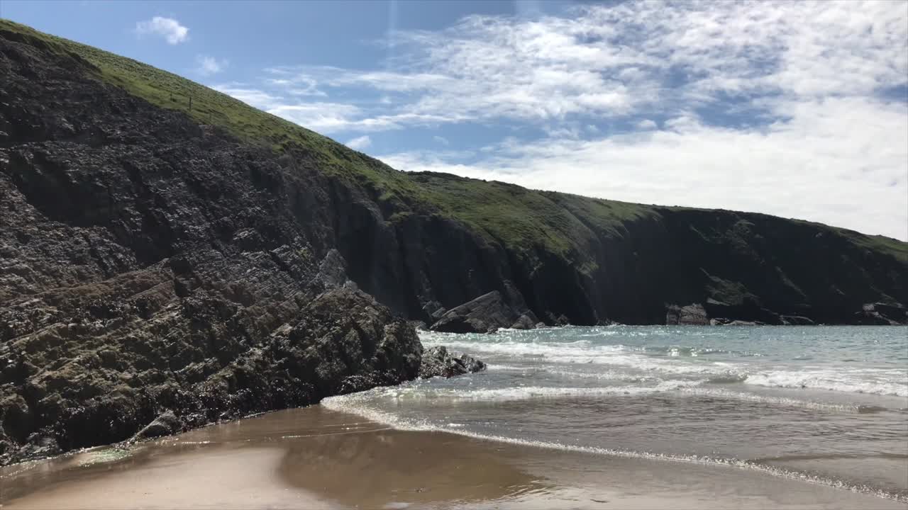 Small waves rolling up sandy beach in front of rugged rocky coastline in Cardigan Bay looming above