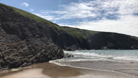 Small-waves-rolling-up-sandy-beach-in-front-of-rugged-rocky-coastline-in-Cardigan-Bay-looming-above