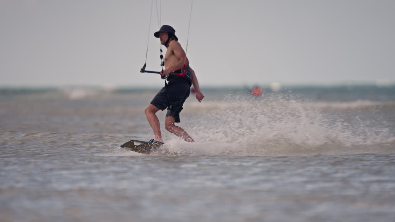 셔츠 없이, 쇼트와 모자를 입고, 긴 회전, 느린 동작을 실행하기 전에 물을 가로질러 날아다니는 카이트보더의 패닝 을 잃습니다.