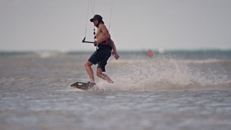 Verlieren-Sie-Sich-In-Einer-Schwenkaufnahme-Eines-Kiteboarders,-Der-Ohne-Hemd-Und-In-Shorts-Und-Mütze-über-Das-Wasser-Gleitet,-Bevor-Er-Eine-Enge-Kurve-Ausführt,-Zeitlupe