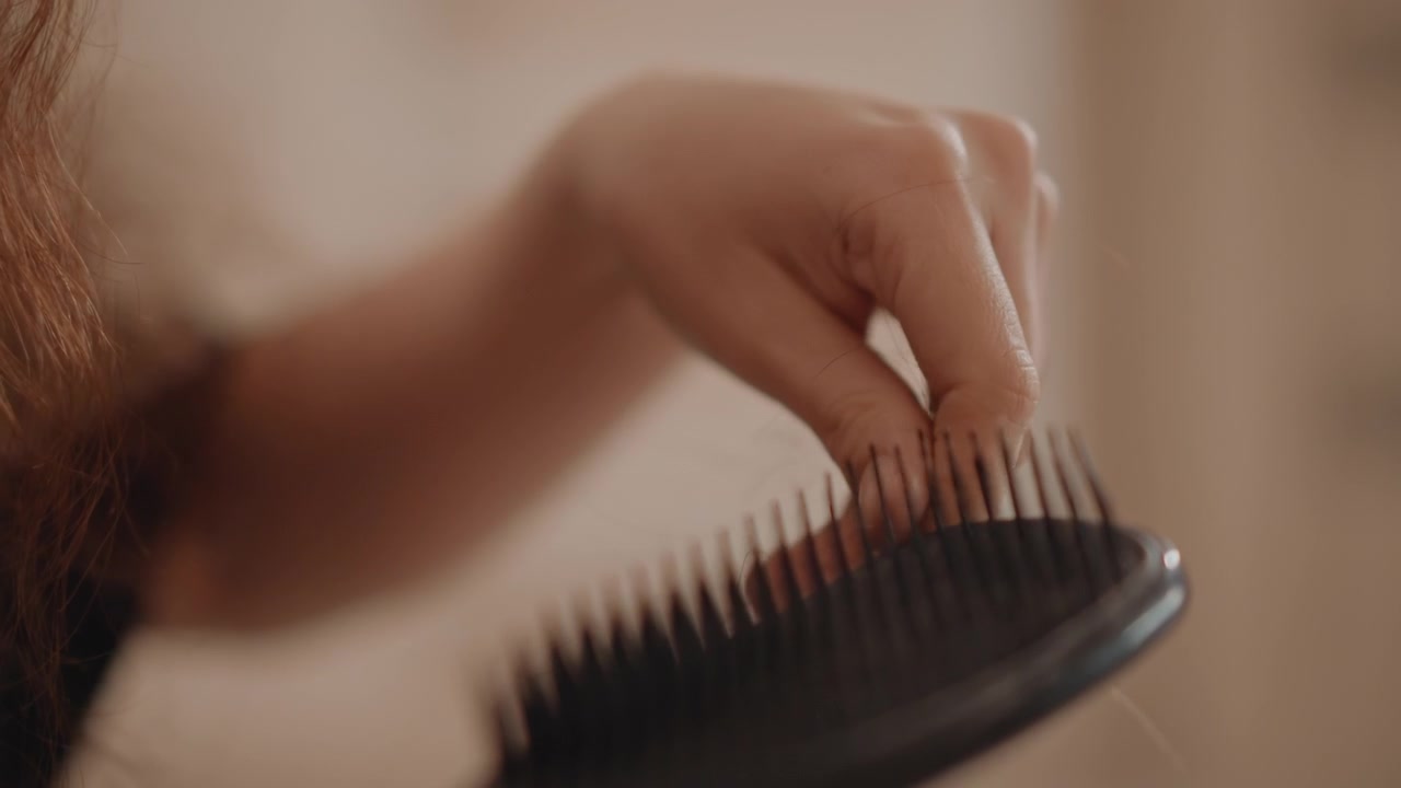 Vista cercana muestra una mano liberando pequeñas gotas hacia un cepillo para el cabello sostenido cerca de cabello fino y claro, con partículas cayendo visibles, cerdas negras flexibles y una acción de aseo tranquila bajo una cálida luz interior.