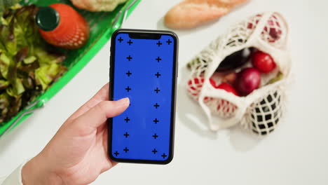 Person holding a smartphone with a blue screen, amidst various groceries