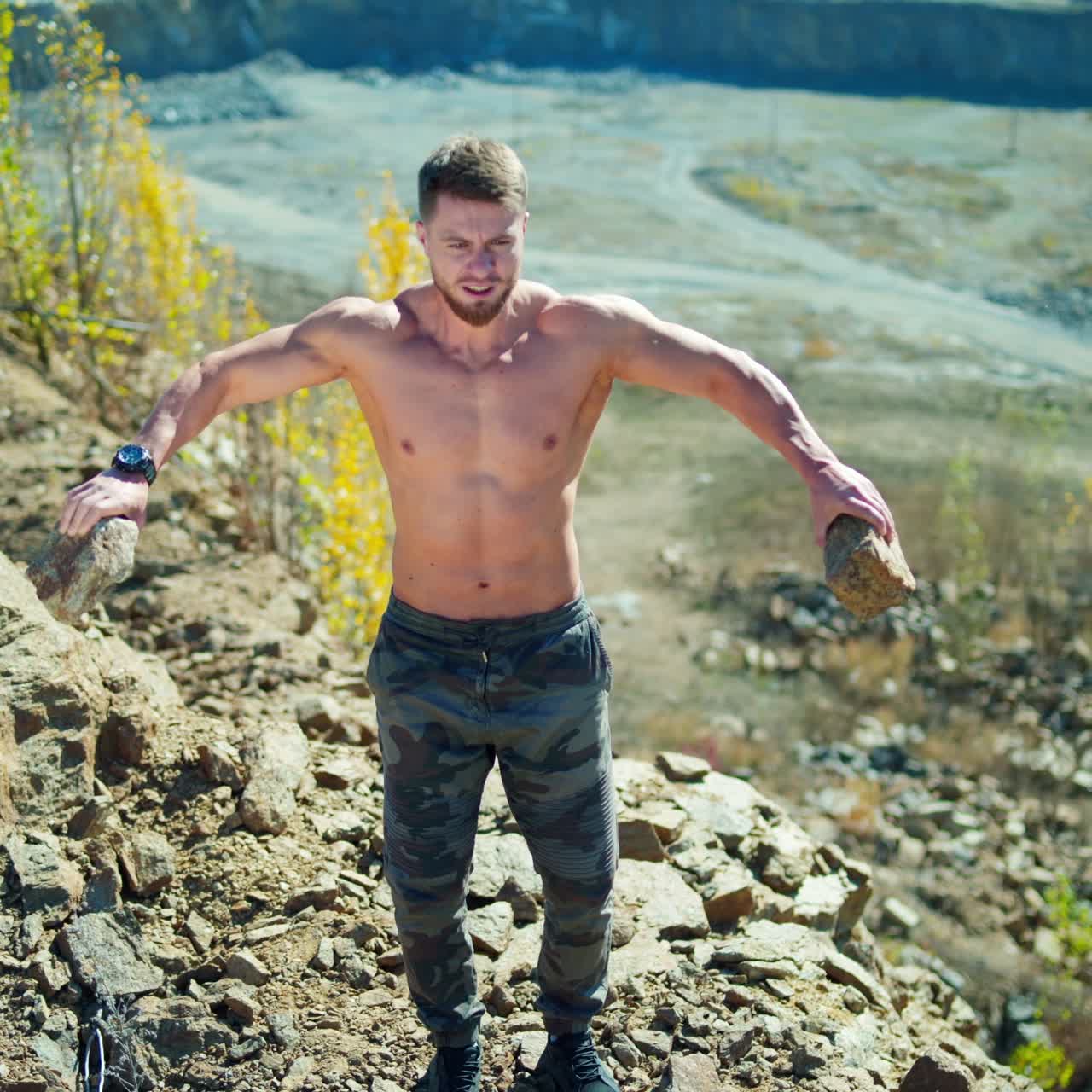 Muscular athlete doing his workout in nature. Shirtless man exercises with stones on the edge of a rocky hill in summer.
