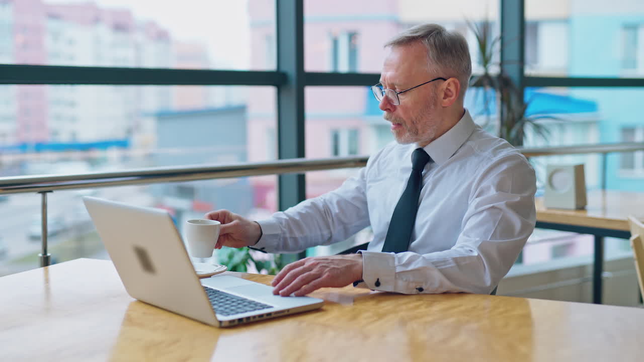 Senior man with a cup working at home on laptop. Freelancer using computer from home-office. Mature businessman working near the window.