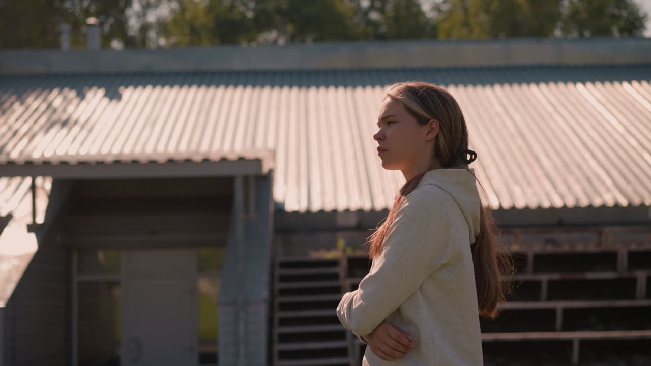 긴 머리카락을 뒤로 고, 후디를 입고, 은 날에 빈 경기장 관중석을 걸어가며, 팔을 접고, 그녀는 깊은 생각에 빠진 것처럼 보이며, 빈 좌석으로 둘러싸여 있습니다.