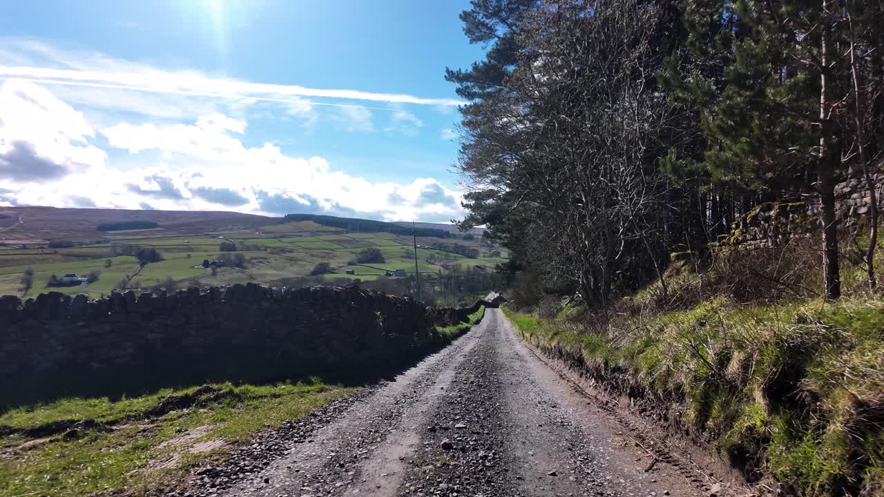 Carretera rural serpenteando a través de colinas verdes con un muro de piedra y bosque bajo un cielo azul. POV, Cámara lenta.