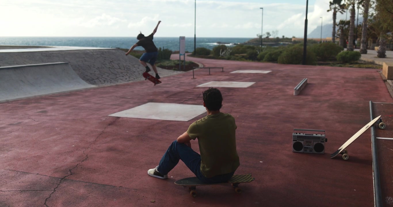 Giovane Seduto sullo Skateboard che Guarda un Amico Eseguire un Trick nello Skatepark Vicino all'Oceano