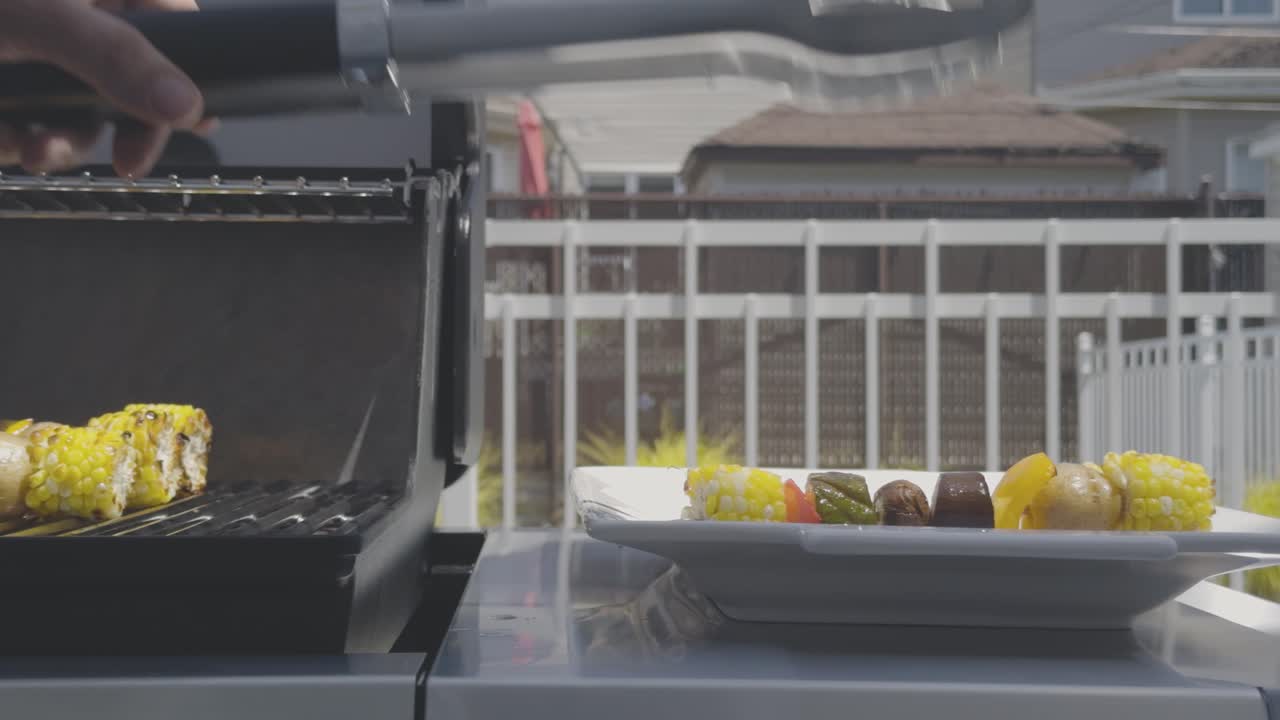 Delicious vegetable skewers with corn, pepper, zucchini, potatoes and mushrooms are cooked on a stainless steel bbq. Juices drip as the tongs pick up one skewer and places it on a plate to the right