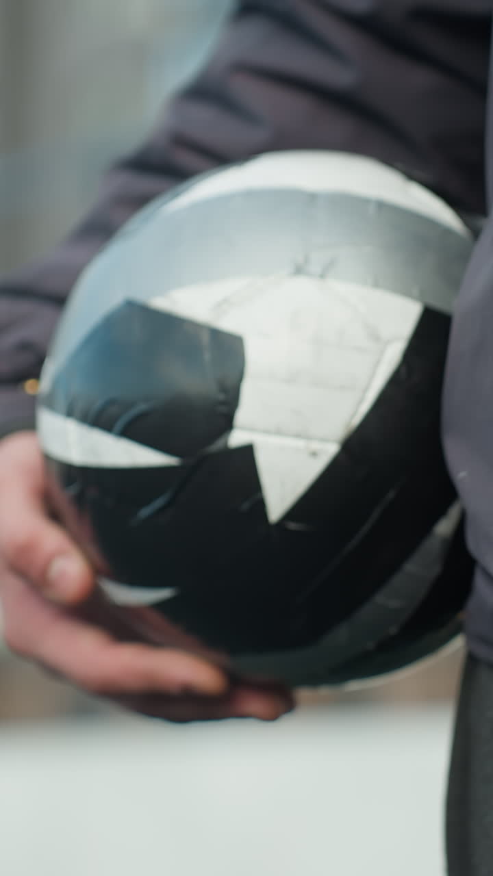 vista lateral de cerca de alguien caminando en un entorno deportivo sosteniendo una pelota de fútbol negra y blanca, mostrando ropa atlética y parte del cuerpo, con fondo urbano