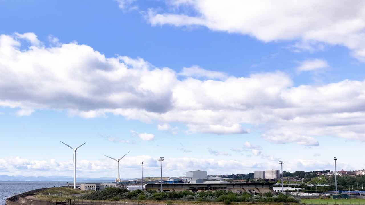 Wide panning shot of wind turbines, industrial buildings, and coastline under bright daylight clouds