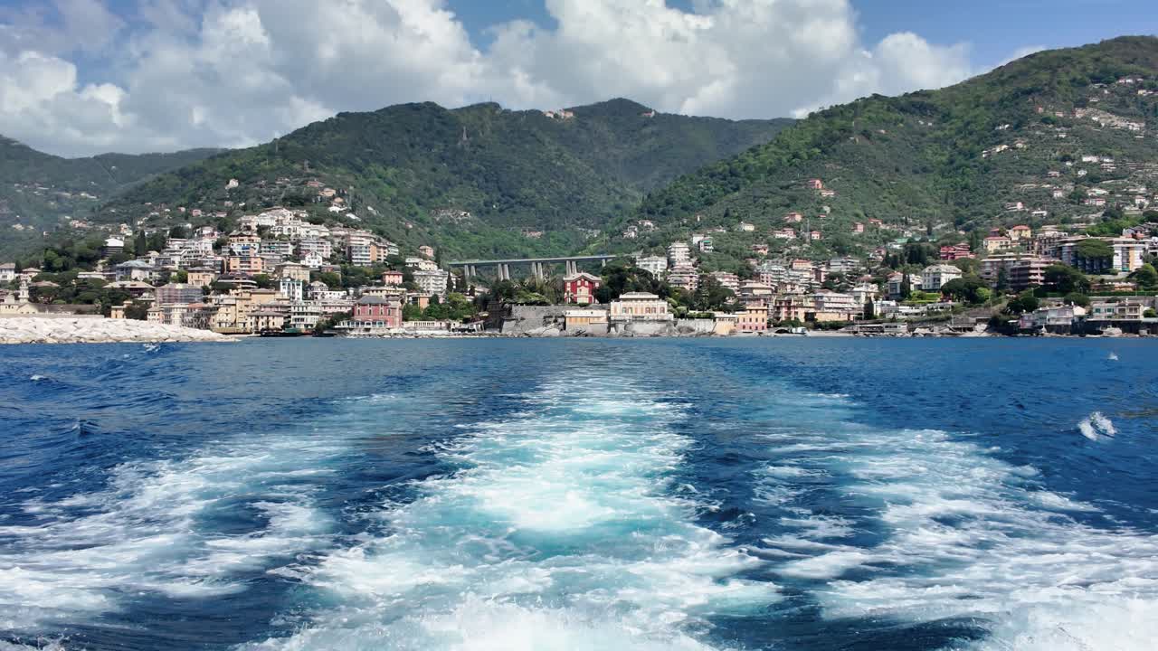 POV dinamico da una barca che si allontana velocemente, con scie sull'acqua blu vibrante, che rivela una pittoresca città costiera incastonata tra montagne lussureggianti, illuminata dal sole sotto cieli vividi, ripresa grandangolare.