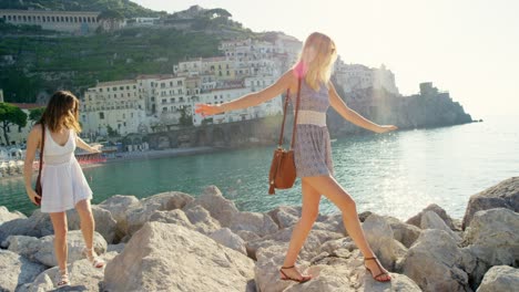 two friends making their way across a series of rocks on the beach. happy women climbing across boulders on the beach