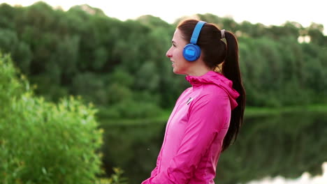 a girl in a pink jacket and blue headphones stands with her back and looks at the river early in the morning after a run