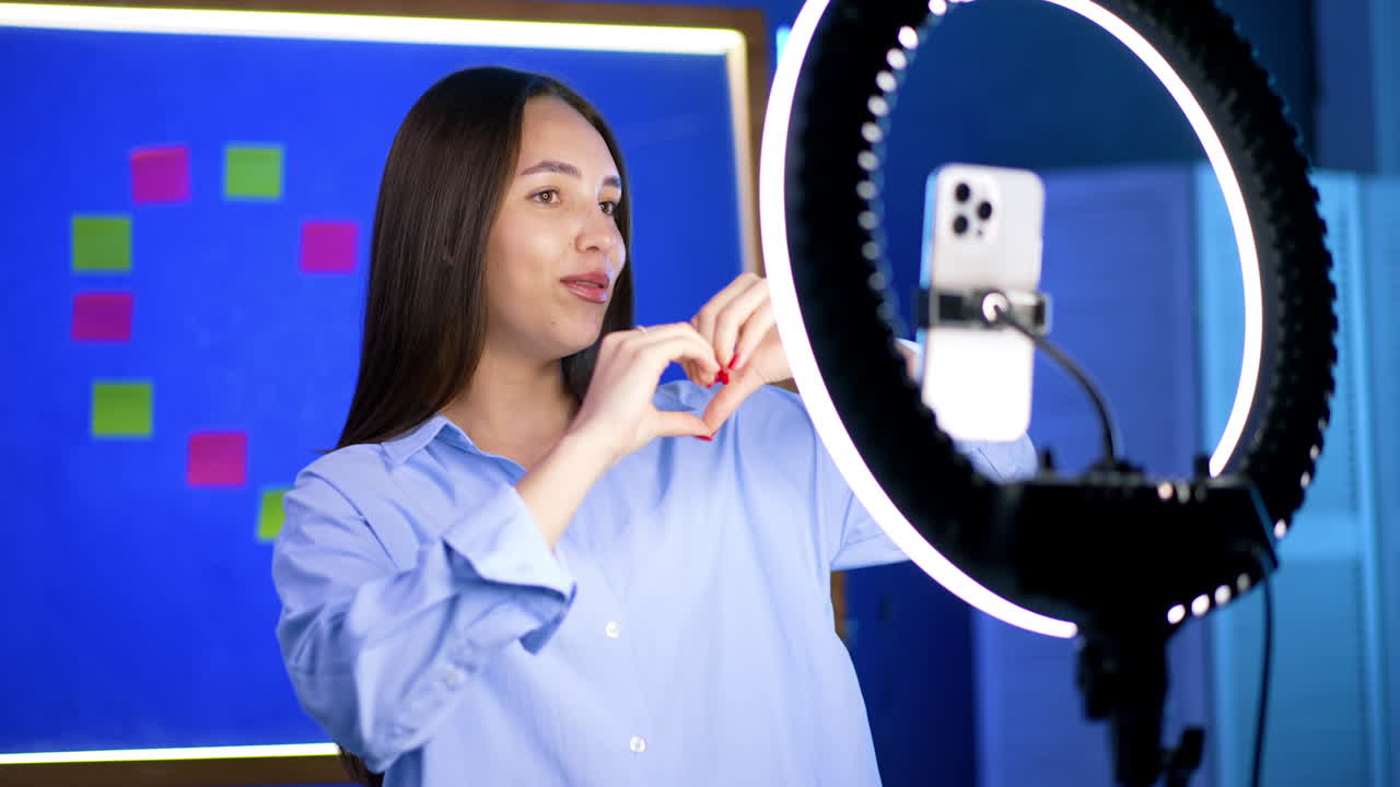 Good-looking Caucasian lady in blue shirt talks to phone camera on tripod with light ring. Girl recording a blog.