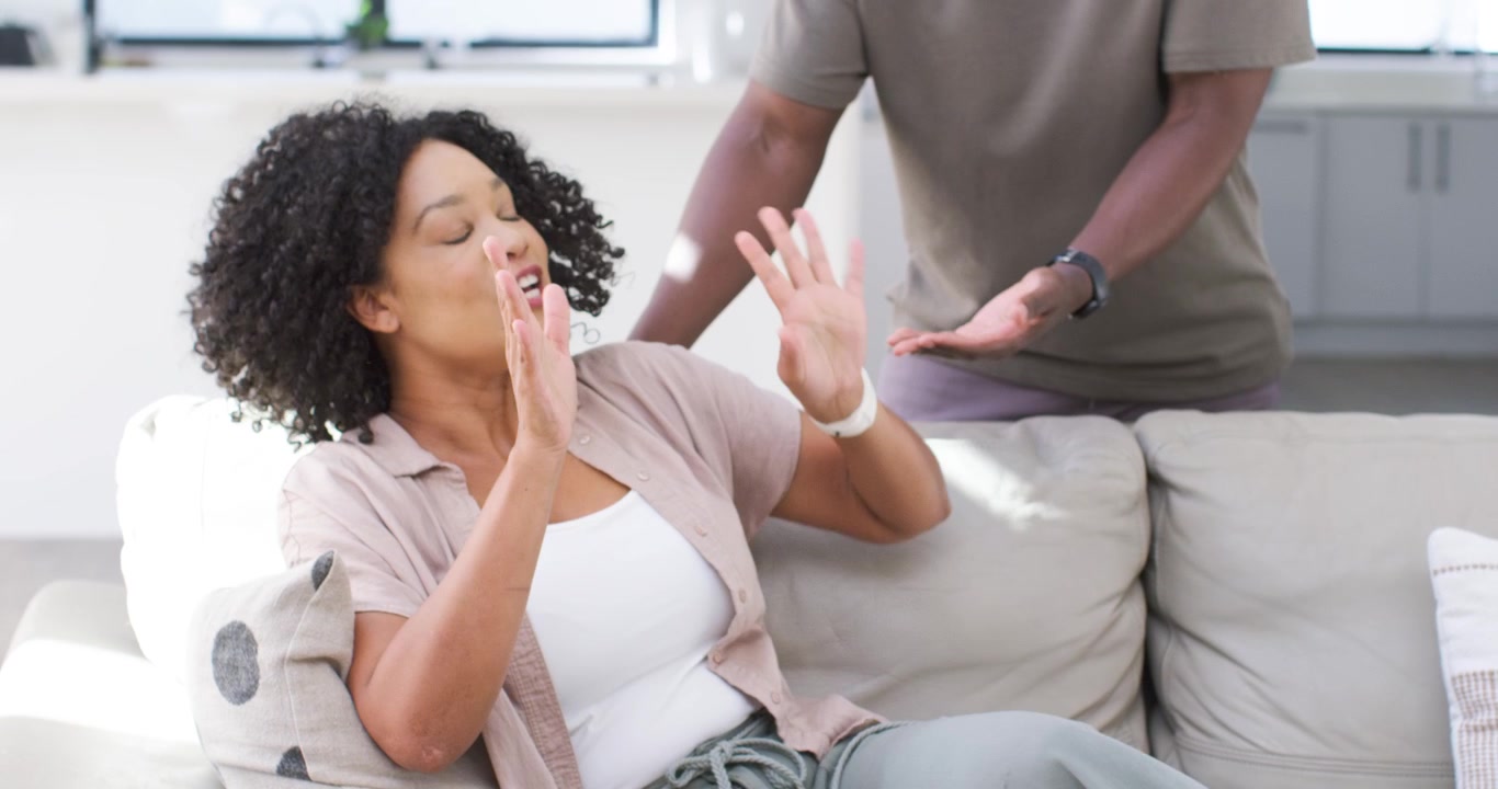 Coppia afroamericana, con la donna che alza le mani sul divano a casa e l'uomo che si avvicina offrendo rassicurazione.