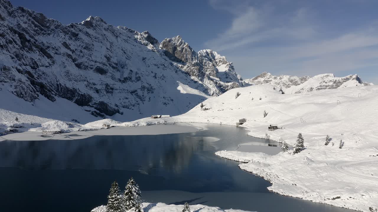 Slow aerial descent over freezing lake towards snow covered trees at Trübsee near Engelberg and Mt. Titlis