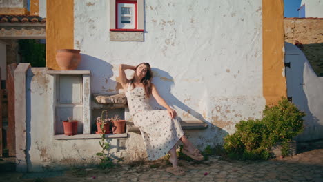 gentle girl warming sunshine leaning house wall. happy lady touching hairstyle