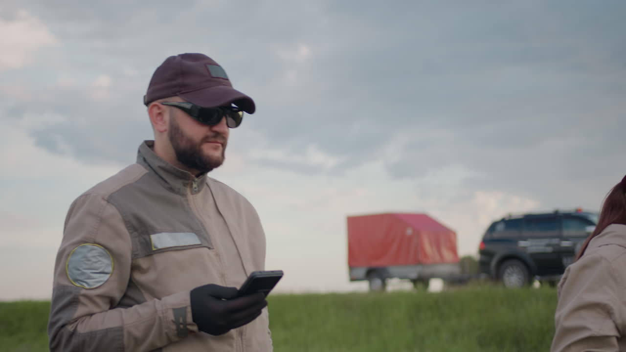 Man in dark cap and sunglasses stands beside woman in gloves as they interact with handheld devices, soft light from blinking car in blurred background under cloudy sky in open grassy roadside field
