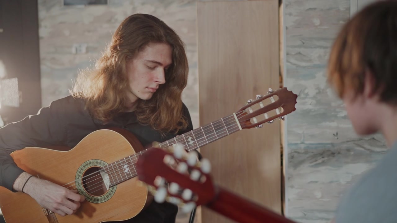 Giovane coach di chitarra caucasico che insegna a uno studente, lezione intima in studio con chitarra acustica, controluce caldo, dimostrazione focalizzata degli accordi, allievo attento che prende appunti, atmosfera di tutoraggio di supporto