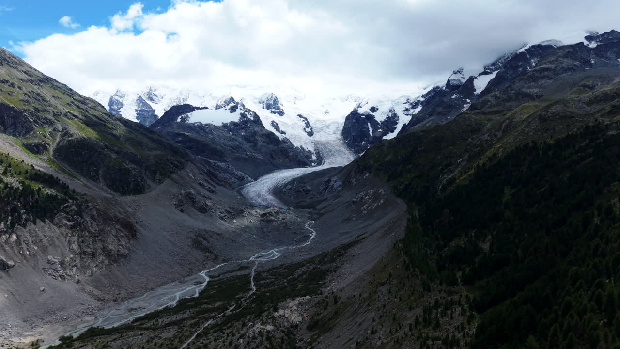 Drone flies forward along a mountain slope toward the glacier river and snowy peaks under cloudy sky